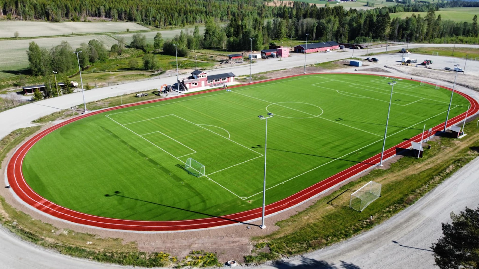 Luftfoto av Bruvollen stadion med kunstgressbane, 400‑meters løpebane rundt og klubbhus med tilhørende bygg, omgitt av skog og jordbrukslandskap.