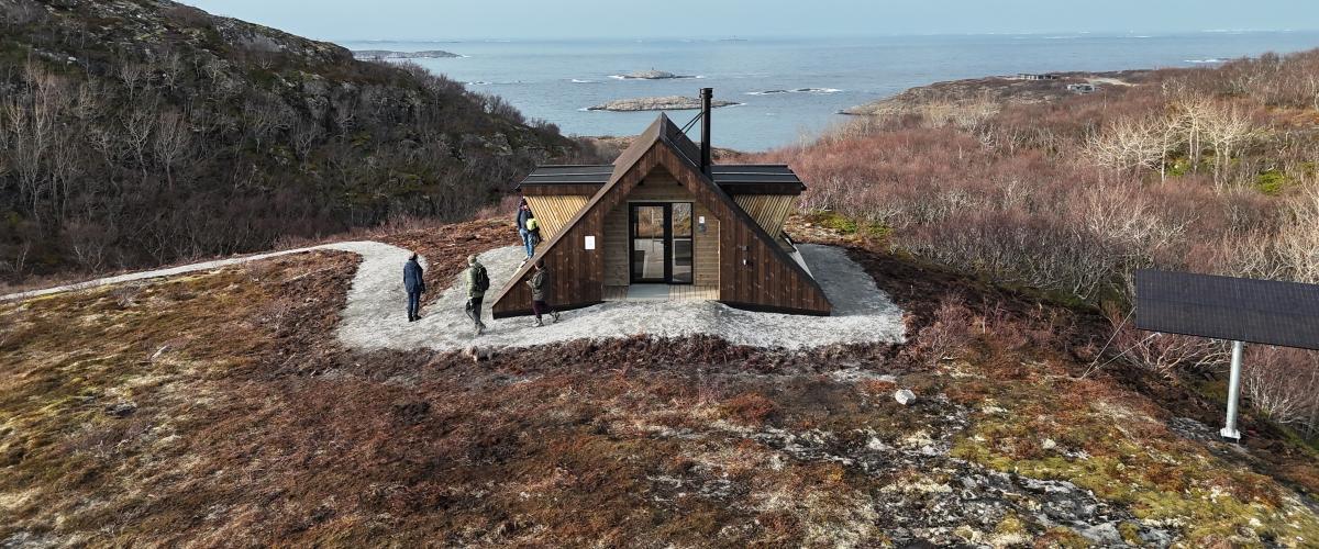 Dagsturhytta ved Utrosjøen ligger på en åpen høyde med utsikt mot hav og holmer, omgitt av karrig kystlandskap, med flere turgåere på stien og et solcellepanel like ved. Foto: Jonas Ali Ghanizadeh