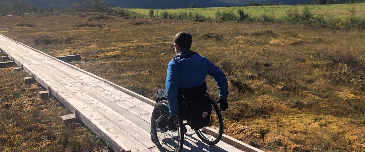 Person i rullestol triller på moderne kavlveg gjennom myrlandskapet.