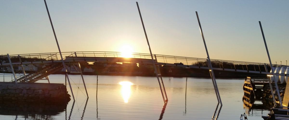 Gangbro med skråstilte pilarer. I bakgrunnen står midnattsola lavt over horisonten.