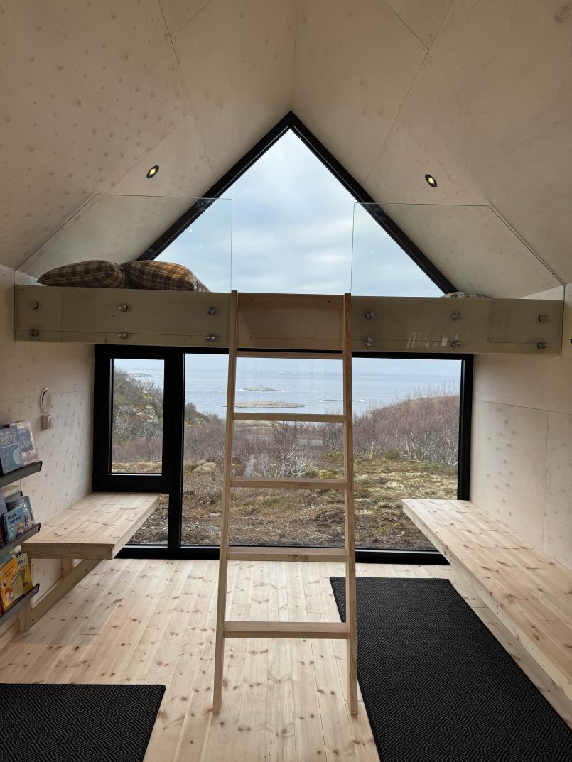 Lyst interiør i dagsturhytta ved Utrosjøen, med store vinduer mot havet, en hems med madrasser og stige opp, benker langs veggene og bokhyller med bøker.
