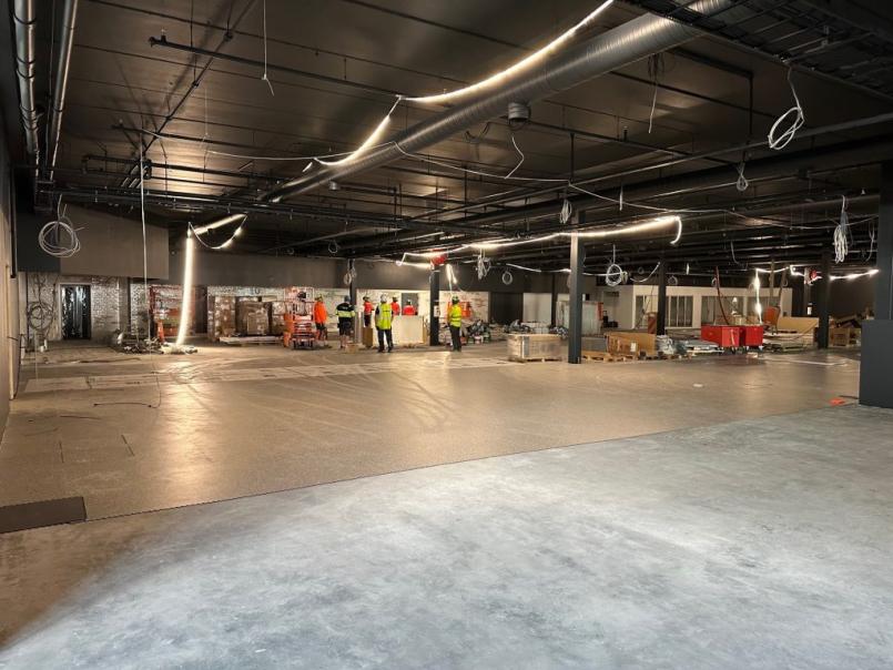 Innendørs byggeplass i dagligvarearealet under isflaten på Eckbobanen, med uferdig gulv, åpne takinstallasjoner og flere byggearbeidere i refleksvester som jobber blant paller og utstyr i bakgrunnen