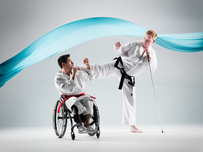 To kampsportutøvere der en sitter i rullestol mens en annen utøver utfører et spark. Utøveren som sparker er stående og blind. Støtter seg derfor på en blindestokk.