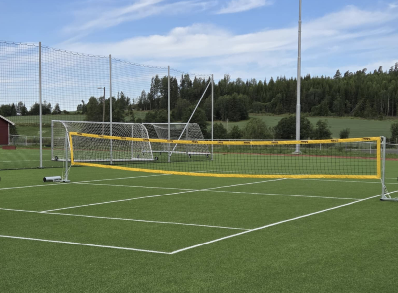 Fotballtennisbane på kunstgresset på Bruvollen, med gule nett spent opp over hvitmerkede baner og fotballmål og ballfangernett i bakgrunnen, omgitt av skog og jordbrukslandskap.