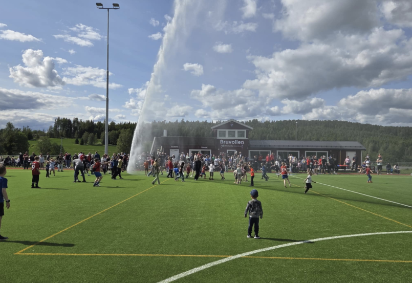 Barn og voksne leker og løper på kunstgressbanen på Bruvollen mens vanningsanlegget skyter en høy vannstråle til værs foran klubbhuset på en solfylt dag.