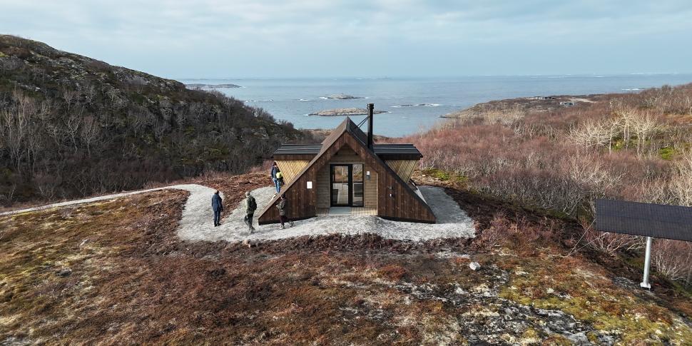Dagsturhytta ved Utrosjøen ligger på en åpen høyde med utsikt mot hav og holmer, omgitt av karrig kystlandskap, med flere turgåere på stien og et solcellepanel like ved. Foto: Jonas Ali Ghanizadeh