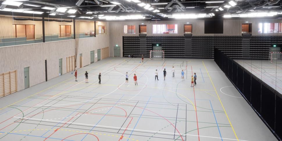 Oversiktsbilde av stor idrettshall med flere håndballbaner og elever i aktivitet. Hallen har teleskoptribune, høyt svart tak med synlige ventilasjonsrør, og store vinduer inn mot møterom og publikumsarealer.